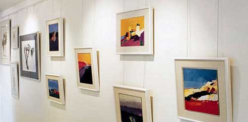 Multiple framed artworks of different sizes hang on a white wall in a gallery setting using a picture hanging system.