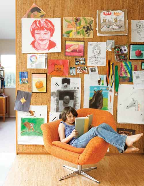 Girl Reading in Front of Art-Covered Cork Wall Young girl reading in an orange chair in front of a cork wall covered with children's artwork and drawings.