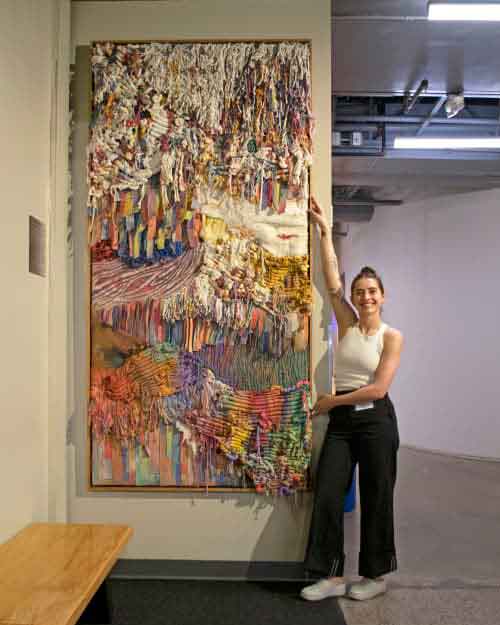 Colorful Textile Artwork Large colorful textile artwork hanging on a wall, with an artist standing beside it, smiling and pointing to the piece.
