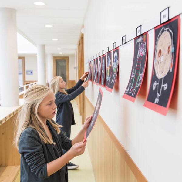 Two school students adjust colourful drawings clipped to a white ArtiTeq Info Rail in a bright corridor. The display rail keeps student artwork neat, easy to swap and protects the walls. Available in Australia from Hang Logic, importer of ArtiTeq picture hanging rails.